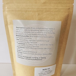 Może przedstawiać: Zamykana torebka z brązowego papieru pakowego zawierająca proszek Moringa. Etykieta zawiera opis, profil botaniczny, zastosowanie, instrukcję przygotowania dla 1-2 g w 200 ml wody, składniki, pochodzenie, przechowywanie i ostrzeżenie. Mała biała etykieta pokazuje MOR-100.