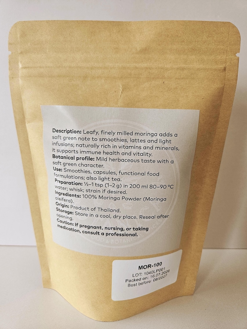 Może przedstawiać: Zamykana torebka z brązowego papieru pakowego zawierająca proszek Moringa. Etykieta zawiera opis, profil botaniczny, zastosowanie, instrukcję przygotowania dla 1-2 g w 200 ml wody, składniki, pochodzenie, przechowywanie i ostrzeżenie. Mała biała etykieta pokazuje MOR-100.