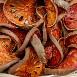 May include: Close-up of dried bael fruit slices, displaying a vibrant orange-red colour with visible seeds. The fruit pieces have a textured surface and are arranged in a woven basket, highlighting their natural form and colour.