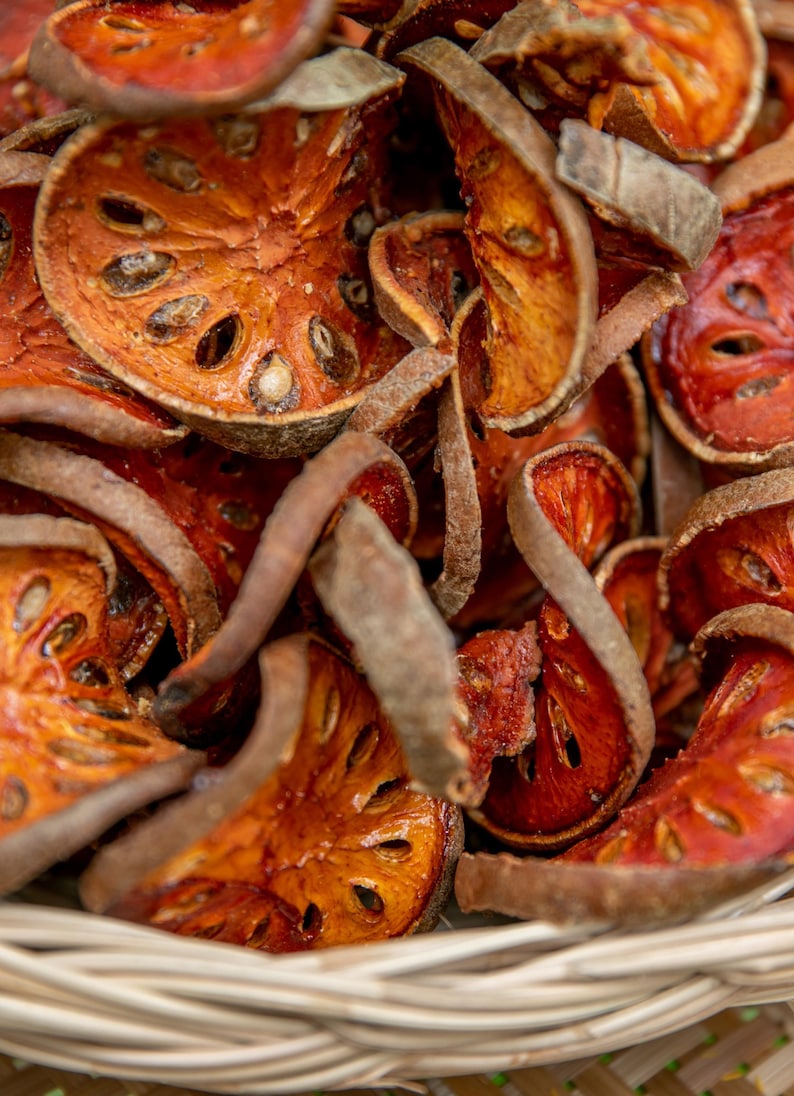 May include: Close-up of dried bael fruit slices, displaying a vibrant orange-red colour with visible seeds. The fruit pieces have a textured surface and are arranged in a woven basket, highlighting their natural form and colour.