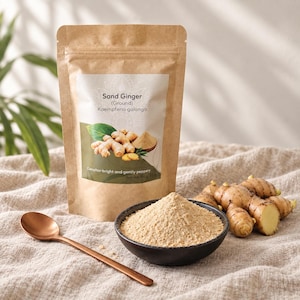 Può includere: Una busta di carta marrone di zenzero aromatico macinato etichettata Kaempferia galanga, accanto a una ciotola nera riempita di polvere beige e un cucchiaio color rame. Una radice di zenzero fresca poggia su un tessuto beige.