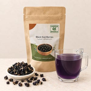 Może przedstawiać: Brązowa papierowa torebka z etykietą Green Botanica Black Goji Berries Lycium ruthenicum o wadze netto 50g, biała miseczka wypełniona suszonymi czarnymi jagodami goji oraz przezroczysty szklany kubek z ciemnofioletowym naparem.