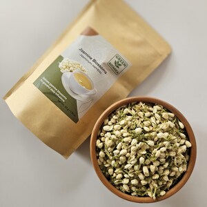 Peut inclure: Un bol en bois rempli de fleurs de jasmin s&eacute;ch&eacute;es &agrave; c&ocirc;t&eacute; d'un sac en papier kraft. Le sac porte une &eacute;tiquette indiquant "Jasmine Blossoms" et "Green Botanica". Les fleurs sont de couleur vert clair.