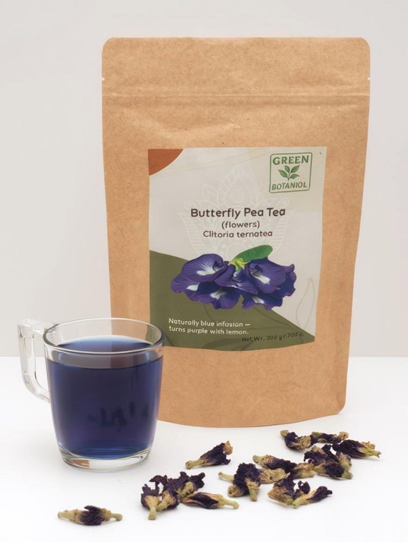 Pode incluir: Um saco de papel castanho com o r&oacute;tulo Green Botaniol Butterfly Pea Tea Clitoria ternatea com um peso l&iacute;quido de 200 g. Ao lado, uma caneca de vidro transparente cheia de l&iacute;quido azul escuro e v&aacute;rias flores secas roxas espalhadas.