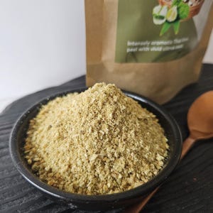 May include: A black bowl containing a light brown, granular spice or seasoning. A copper spoon sits beside the bowl on a dark wooden surface. A brown paper bag with text and a green label is in the background.