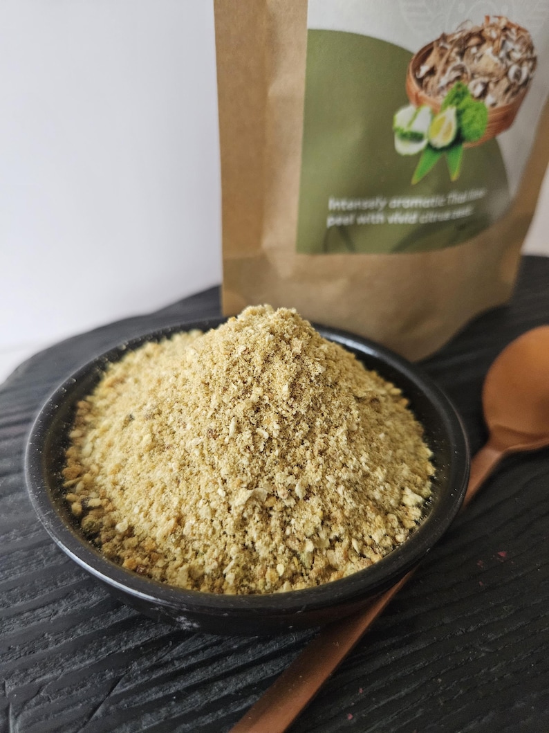 May include: A black bowl containing a light brown, granular spice or seasoning. A copper spoon sits beside the bowl on a dark wooden surface. A brown paper bag with text and a green label is in the background.