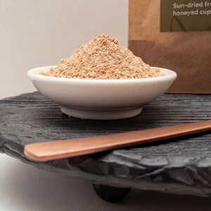 May include: A white ceramic bowl filled with light brown powder sits on a black, textured wooden tray. A copper-coloured spoon rests on the tray. A brown paper bag with text is in the background. The image suggests a food or spice product.