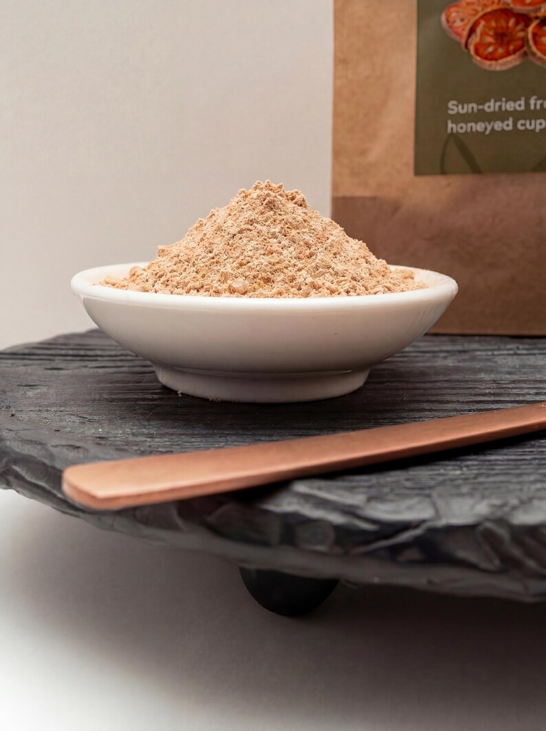 May include: A white ceramic bowl filled with light brown powder sits on a black, textured wooden tray. A copper-coloured spoon rests on the tray. A brown paper bag with text is in the background. The image suggests a food or spice product.