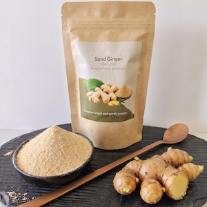 Op de afbeelding: Een bruine papieren zak met het label Sand Ginger (Ground) Kaempferia galanga, met 100g specerij. Daarnaast staat een zwarte kom gevuld met lichtbruin gemberpoeder, een lange houten lepel en een verse, lichtbruine gemberwortelstok.