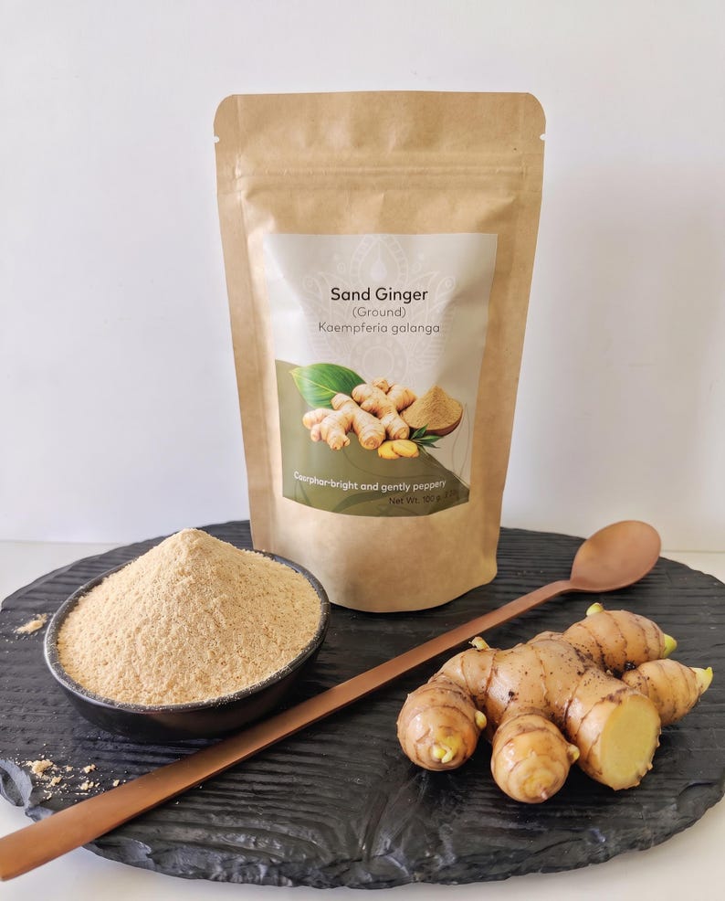Op de afbeelding: Een bruine papieren zak met het label Sand Ginger (Ground) Kaempferia galanga, met 100g specerij. Daarnaast staat een zwarte kom gevuld met lichtbruin gemberpoeder, een lange houten lepel en een verse, lichtbruine gemberwortelstok.