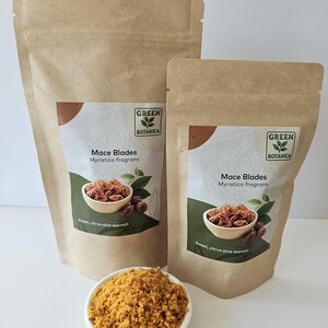 K&ouml;nnte beinhalten: Zwei braune Papierbeutel mit der Aufschrift Green Botanica Mace Blades Myristica fragrans und einem Bild von Muskatbl&uuml;ten in einer Schale. Eine kleine wei&szlig;e Schale mit gemahlenem gelb-orangem Gew&uuml;rz steht vor den Beuteln auf einer wei&szlig;en Fl&auml;che.