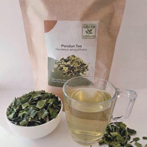 Op de afbeelding: Een heldere glazen mok gevuld met lichtgroene thee staat naast een witte kom met groene pandanbladeren. Een papieren zak met Pandan Tea staat achter de mok en kom. De zak heeft de tekst "GREEN BOTANICA" en "Pandan Tea Pandanus amaryllifolius".