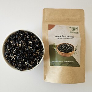 K&ouml;nnte beinhalten: Eine wei&szlig;e Schale gef&uuml;llt mit getrockneten schwarzen Goji-Beeren neben einem braunen Papierbeutel mit der Aufschrift Green Botanica Black Goji Berries Lycium ruthenicum, der nat&uuml;rlich blau-violett mit Zitrone wird, Nettogewicht 50 Gramm.