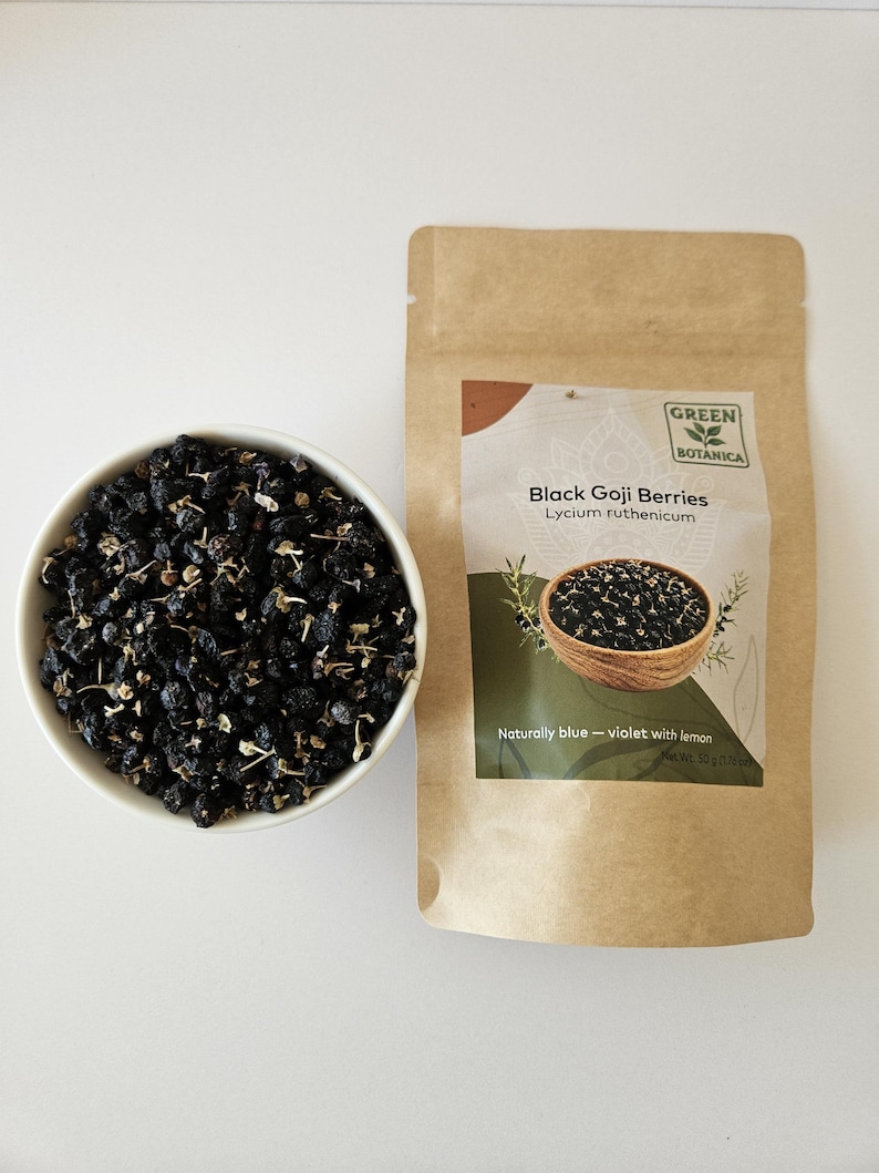 K&ouml;nnte beinhalten: Eine wei&szlig;e Schale gef&uuml;llt mit getrockneten schwarzen Goji-Beeren neben einem braunen Papierbeutel mit der Aufschrift Green Botanica Black Goji Berries Lycium ruthenicum, der nat&uuml;rlich blau-violett mit Zitrone wird, Nettogewicht 50 Gramm.