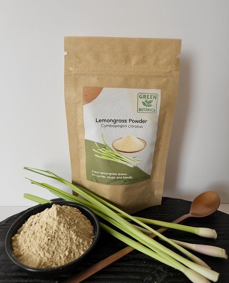 May include: A brown paper bag of lemongrass powder with a green and white label that reads "Lemongrass Powder". A black bowl of lemongrass powder, fresh lemongrass stalks, and a wooden spoon are in the foreground. The product is from Green Botanica.