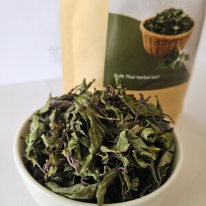 K&ouml;nnte beinhalten: Eine wei&szlig;e Sch&uuml;ssel gef&uuml;llt mit getrockneten gr&uuml;nen Bl&auml;ttern und violetten St&auml;ngeln. Dahinter steht ein beigefarbener Beutel mit der Aufschrift Java Tea (Cat's Whiskers) Orthosiphon aristatus, einem Bild eines Bl&auml;tterkorbs und dem Text Soft Thai herbal leaf.