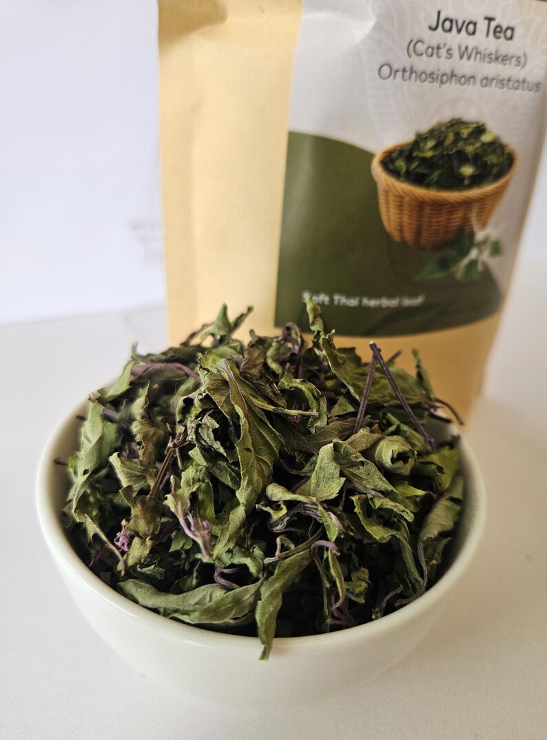 K&ouml;nnte beinhalten: Eine wei&szlig;e Sch&uuml;ssel gef&uuml;llt mit getrockneten gr&uuml;nen Bl&auml;ttern und violetten St&auml;ngeln. Dahinter steht ein beigefarbener Beutel mit der Aufschrift Java Tea (Cat's Whiskers) Orthosiphon aristatus, einem Bild eines Bl&auml;tterkorbs und dem Text Soft Thai herbal leaf.