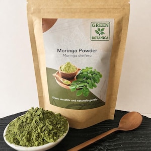 Może przedstawiać: Brązowa papierowa torebka z proszkiem Moringa marki Green Botanica z etykietą Moringa oleifera i wagą netto 100 g. Mała biała miseczka wypełniona zielonym proszkiem oraz drewniana łyżka znajdują się przed torebką na ciemnej powierzchni.