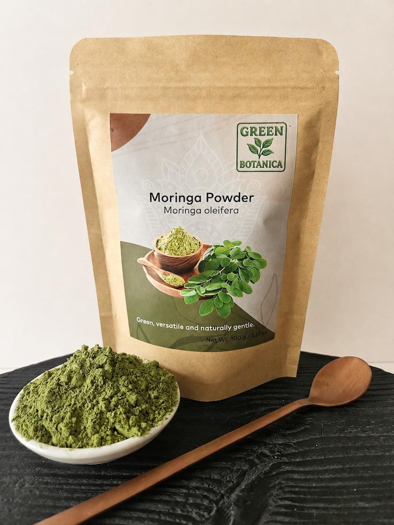 Może przedstawiać: Brązowa papierowa torebka z proszkiem Moringa marki Green Botanica z etykietą Moringa oleifera i wagą netto 100 g. Mała biała miseczka wypełniona zielonym proszkiem oraz drewniana łyżka znajdują się przed torebką na ciemnej powierzchni.