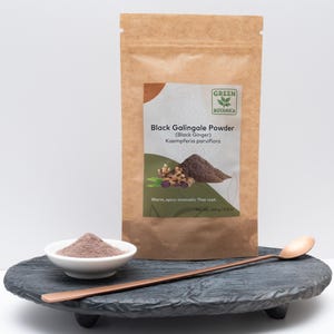 Op de afbeelding: Een bruine papieren zak met zwarte galangalpoeder, met de tekst "Green Botanica" en "Black Galingale Powder (Black Ginger)". Een kleine witte kom met poeder en een koperen lepel liggen op een donkergrijs, verhoogd platform.