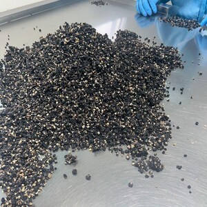 K&ouml;nnte beinhalten: Ein gro&szlig;er Haufen kleiner, getrockneter schwarzer Goji-Beeren mit sichtbaren hellen Stielen, die auf einer glatten, reflektierenden Edelstahloberfl&auml;che liegen. Die Beeren sind unregelm&auml;&szlig;ig geformt, dunkel und wirken dehydriert und schrumpelig.