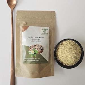 May include: A still life image shows a copper spoon, a brown paper packet, and a small black bowl. The packet is labelled "Kaffir Lime Rinds (ground)" with a "Green Botanica" logo. The bowl contains a light green powder, and the spoon is long and slender.