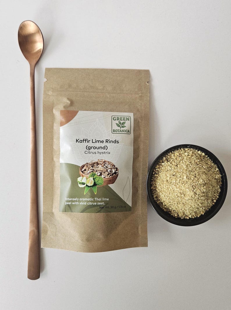 May include: A still life image shows a copper spoon, a brown paper packet, and a small black bowl. The packet is labelled "Kaffir Lime Rinds (ground)" with a "Green Botanica" logo. The bowl contains a light green powder, and the spoon is long and slender.