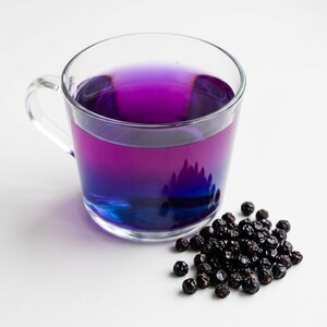 K&ouml;nnte beinhalten: Eine durchsichtige Glastasse gef&uuml;llt mit einer leuchtend violetten Fl&uuml;ssigkeit, die am Boden in ein tiefes Blau &uuml;bergeht. Neben der Tasse liegt ein kleiner, verstreuter Haufen dunkler, getrockneter, runder Beeren mit faltiger Textur.