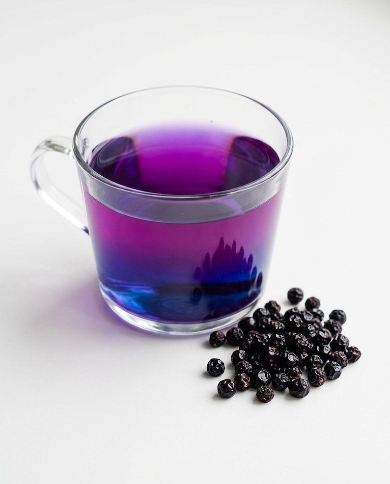 K&ouml;nnte beinhalten: Eine durchsichtige Glastasse gef&uuml;llt mit einer leuchtend violetten Fl&uuml;ssigkeit, die am Boden in ein tiefes Blau &uuml;bergeht. Neben der Tasse liegt ein kleiner, verstreuter Haufen dunkler, getrockneter, runder Beeren mit faltiger Textur.