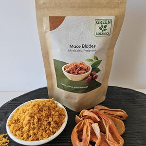 K&ouml;nnte beinhalten: Ein brauner Papierbeutel mit der Aufschrift Green Botanica Mace Blades Myristica fragrans, eine wei&szlig;e Schale mit goldgelbem gemahlenem Muskatbl&uuml;tenpulver und ein kleiner Haufen getrockneter orangefarbener Muskatbl&uuml;ten. Das Etikett zeigt Muskatbl&uuml;ten.