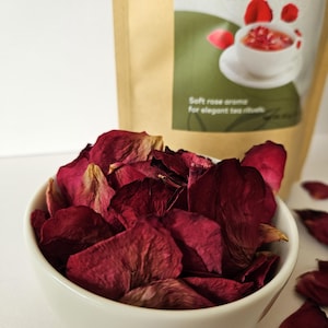 May include: A white bowl filled with dried, deep red rose petals. In the background, a brown paper bag displays the text "Rose Petals Tea" and an illustration of tea. The image suggests a product for tea rituals.