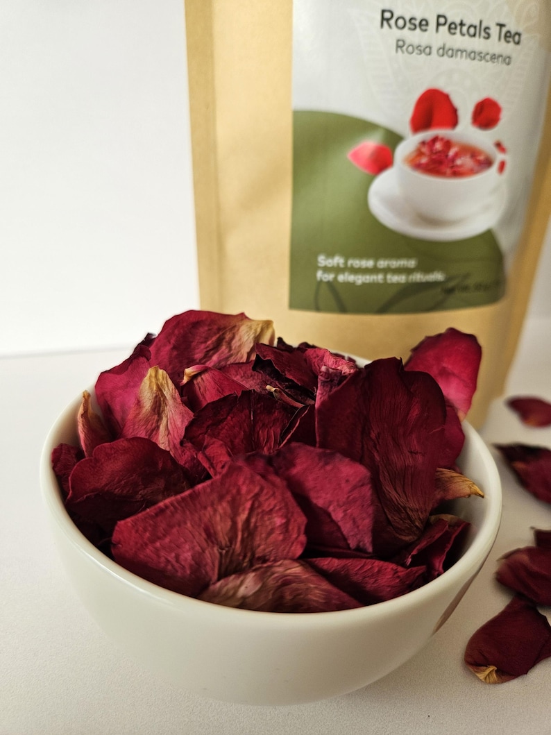 May include: A white bowl filled with dried, deep red rose petals. In the background, a brown paper bag displays the text "Rose Petals Tea" and an illustration of tea. The image suggests a product for tea rituals.
