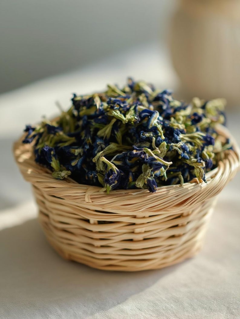 Pode incluir: Uma pequena cesta redonda de vime tran&ccedil;ado em tom castanho claro, cheia com um monte de flores de feij&atilde;o-borboleta secas. As flores s&atilde;o de um azul profundo com caules e c&aacute;lices verde-p&aacute;lidos, empilhadas densamente acima da borda.