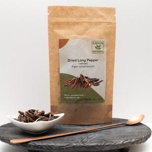 Puede incluir: Una bolsa de papel marrón de pimienta larga seca con el texto "Dried Long Pepper (whole) Piper retrofractum". Un pequeño cuenco blanco de pimienta larga seca y una cuchara de cobre están sobre una bandeja de madera negra y redonda.