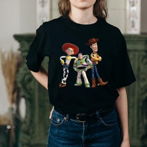 May include: Black t-shirt featuring a colorful graphic of the Toy Story characters Woody, Buzz Lightyear, and Jessie. The design includes the characters in their signature outfits. The shirt is made of a soft material.