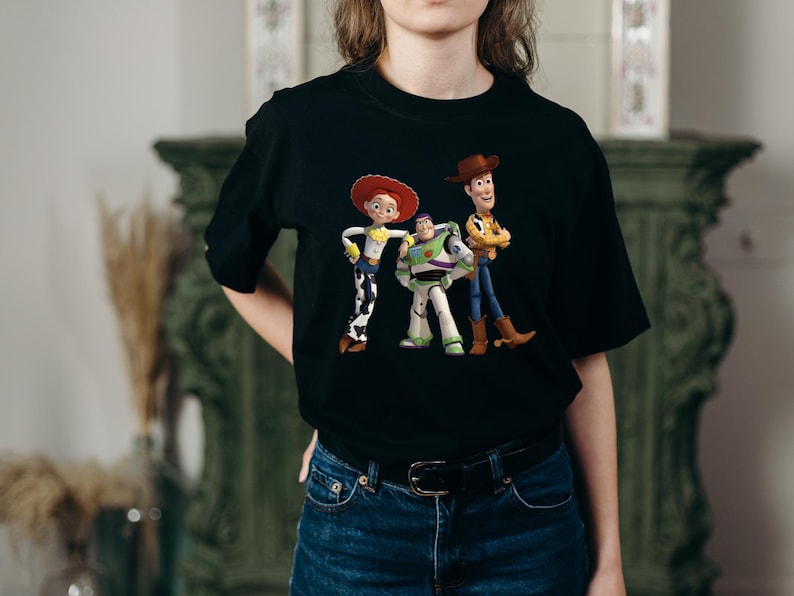 May include: Black t-shirt featuring a colorful graphic of the Toy Story characters Woody, Buzz Lightyear, and Jessie. The design includes the characters in their signature outfits. The shirt is made of a soft material.