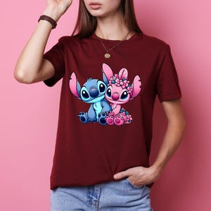 May include: A maroon t-shirt with a cartoon illustration of two characters. One is blue with large ears, the other pink with a floral crown. The graphic is centered on the shirt.