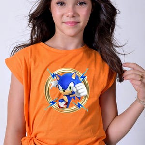 May include: Orange t-shirt featuring a graphic of Sonic the Hedgehog inside a gold ring with blue lightning bolts. The blue hedgehog is in a running pose. The shirt is made of a soft material.