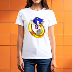 May include: White t-shirt featuring a graphic of Sonic the Hedgehog, a blue anthropomorphic hedgehog, jumping through a golden ring. The shirt is made of a soft material and is suitable for casual wear.