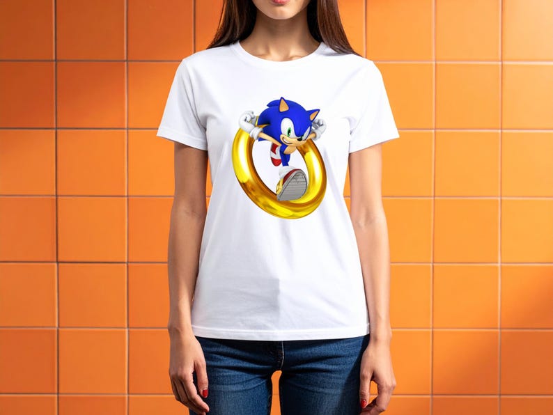 May include: White t-shirt featuring a graphic of Sonic the Hedgehog, a blue anthropomorphic hedgehog, jumping through a golden ring. The shirt is made of a soft material and is suitable for casual wear.