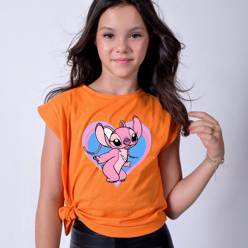 May include: Bright orange t-shirt with a pink cartoon character inside a heart-shaped design with blue and purple accents. The t-shirt has short sleeves and a knotted side detail. The character has large ears and a friendly expression.