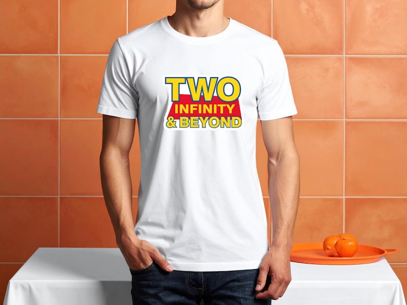 May include: White t-shirt with the text "TWO INFINITY & BEYOND" in yellow and red. The shirt is being worn by a person. The person is wearing blue jeans. Two oranges are on an orange tray.