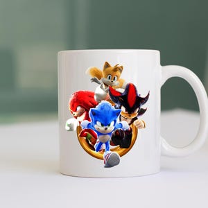 May include: White ceramic mug featuring a colorful graphic of Sonic the Hedgehog and his friends. The design includes Sonic, Tails, Knuckles, and Shadow, set against a gold ring. The mug has a curved handle for easy gripping.
