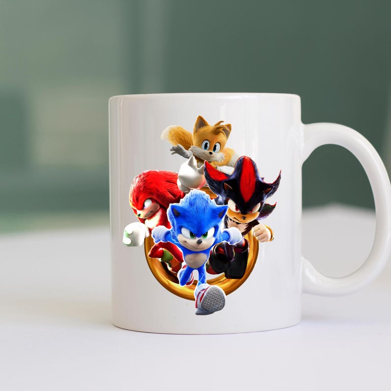 May include: White ceramic mug featuring a colorful graphic of Sonic the Hedgehog and his friends. The design includes Sonic, Tails, Knuckles, and Shadow, set against a gold ring. The mug has a curved handle for easy gripping.
