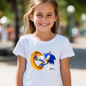 May include: White t-shirt featuring a graphic of Sonic the Hedgehog, a blue anthropomorphic hedgehog, leaping through a golden ring. The design is on a white t-shirt.