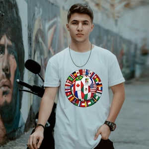 May include: A white short-sleeved t-shirt featuring a central graphic of a soccer ball composed of various national flags. The ball design includes a central panel with the text 2026 and a surrounding ring of smaller international flag icons.