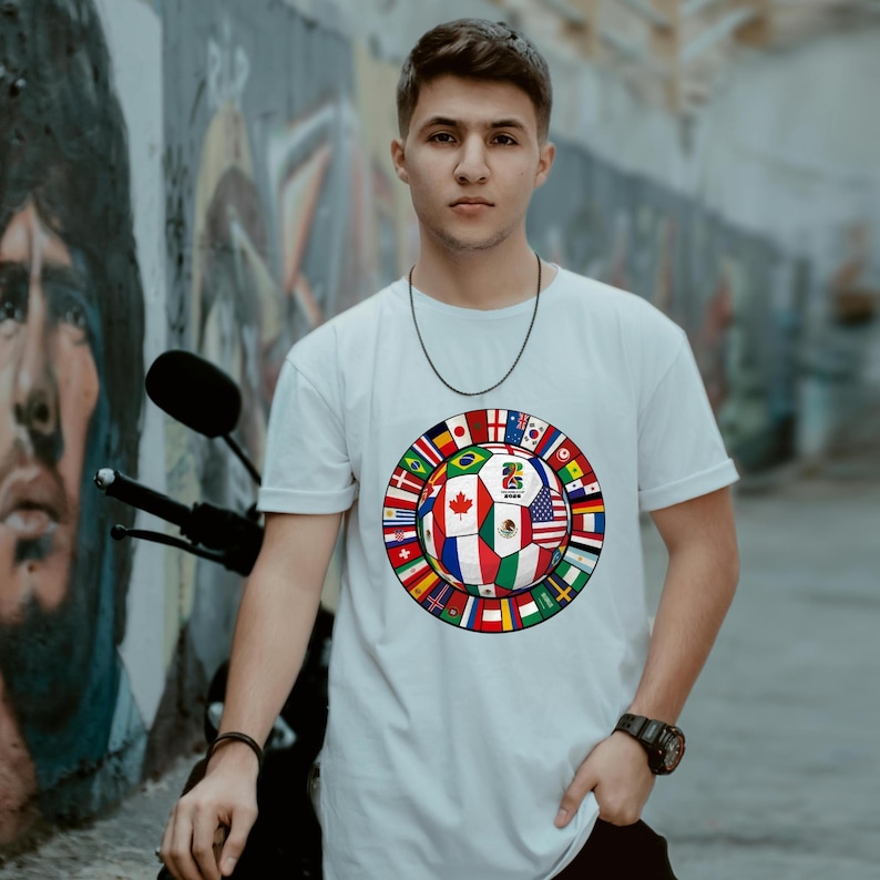 May include: A white short-sleeved t-shirt featuring a central graphic of a soccer ball composed of various national flags. The ball design includes a central panel with the text 2026 and a surrounding ring of smaller international flag icons.
