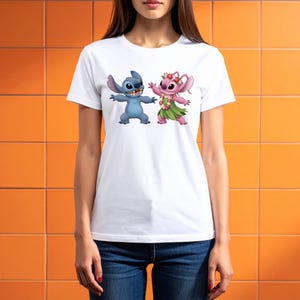 May include: White t-shirt with a cartoon image of Stitch and Angel. Stitch is blue, Angel is pink, both with arms outstretched. Angel wears a flower crown and grass skirt. The t-shirt has a classic crew neck.