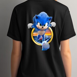 May include: Black t-shirt featuring a vibrant graphic of Sonic the Hedgehog in a running pose. The character is depicted in blue with white gloves and shoes, set against a sunset scene within a golden ring. A fun, eye-catching design for fans.
