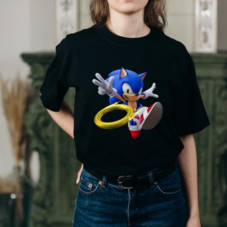 May include: Black t-shirt featuring a vibrant graphic of Sonic the Hedgehog, a blue anthropomorphic hedgehog, with a gold ring. Sonic is depicted mid-jump with a peace sign. The shirt is made of a soft material.